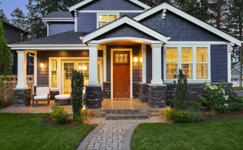 Modern two-story home with dark blue exterior, white columns, warm-lit windows, red front door, stone accents, brick pathway, and manicured lawn at dusk