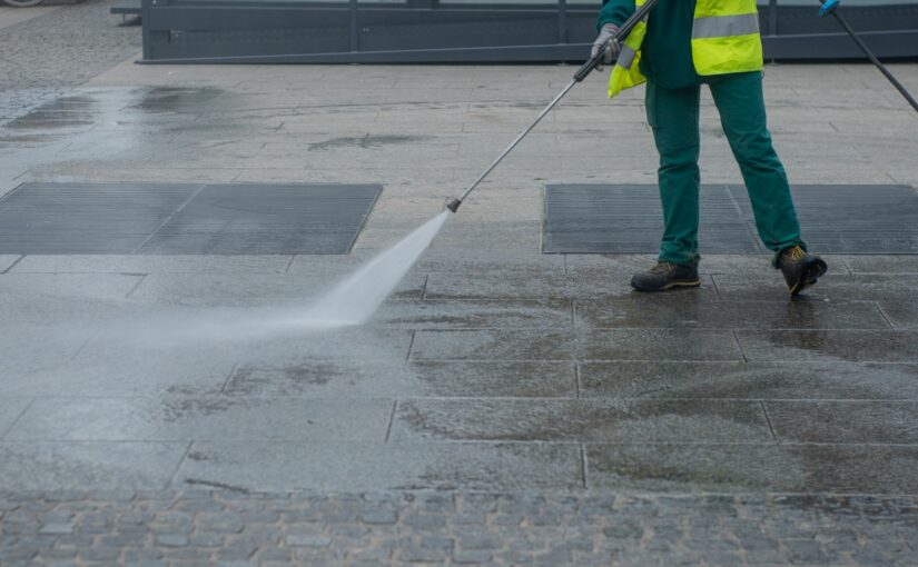 Street cleaner in high-visibility vest using pressure washer to clean wet pavement in urban setting